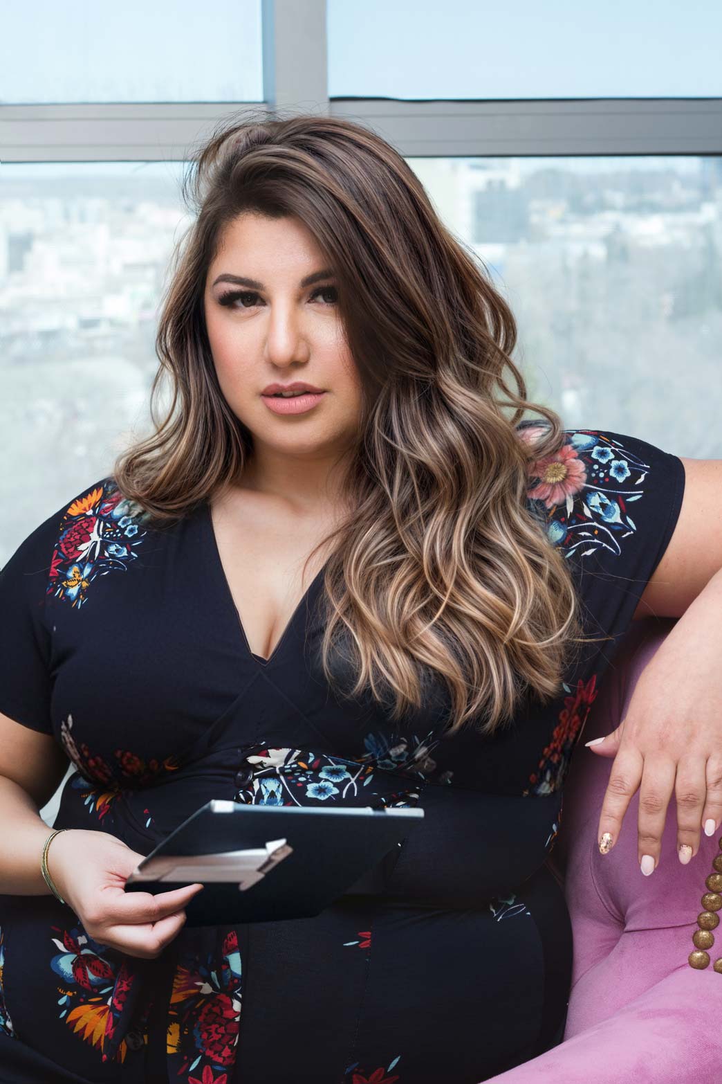 Retrato de Daiana, CEO de DR Estética, sentada en un sillón rosa, sosteniendo una tablet. Viste un vestido negro con estampados florales, con un fondo de ventana que muestra una vista urbana.