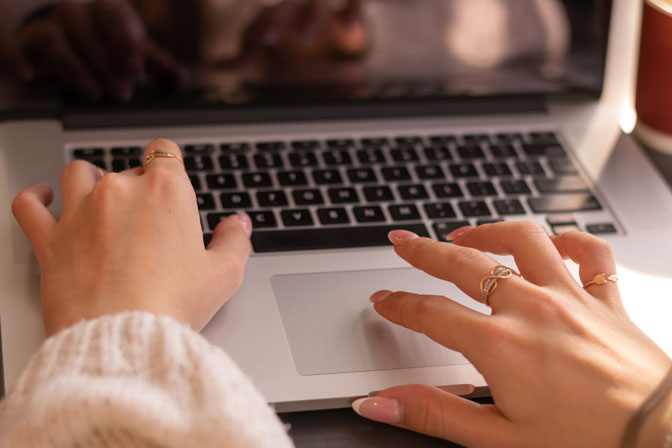 Manos femeninas escribiendo en el teclado de un portátil, con anillos dorados y uñas cuidadas, en un ambiente cálido y profesional.